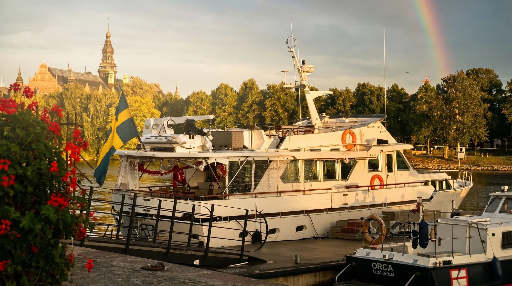 M/Y Gaia vid Strandvägen i Stockholm — regnbåge över hamnen med Nordiska museet i bakgrunden